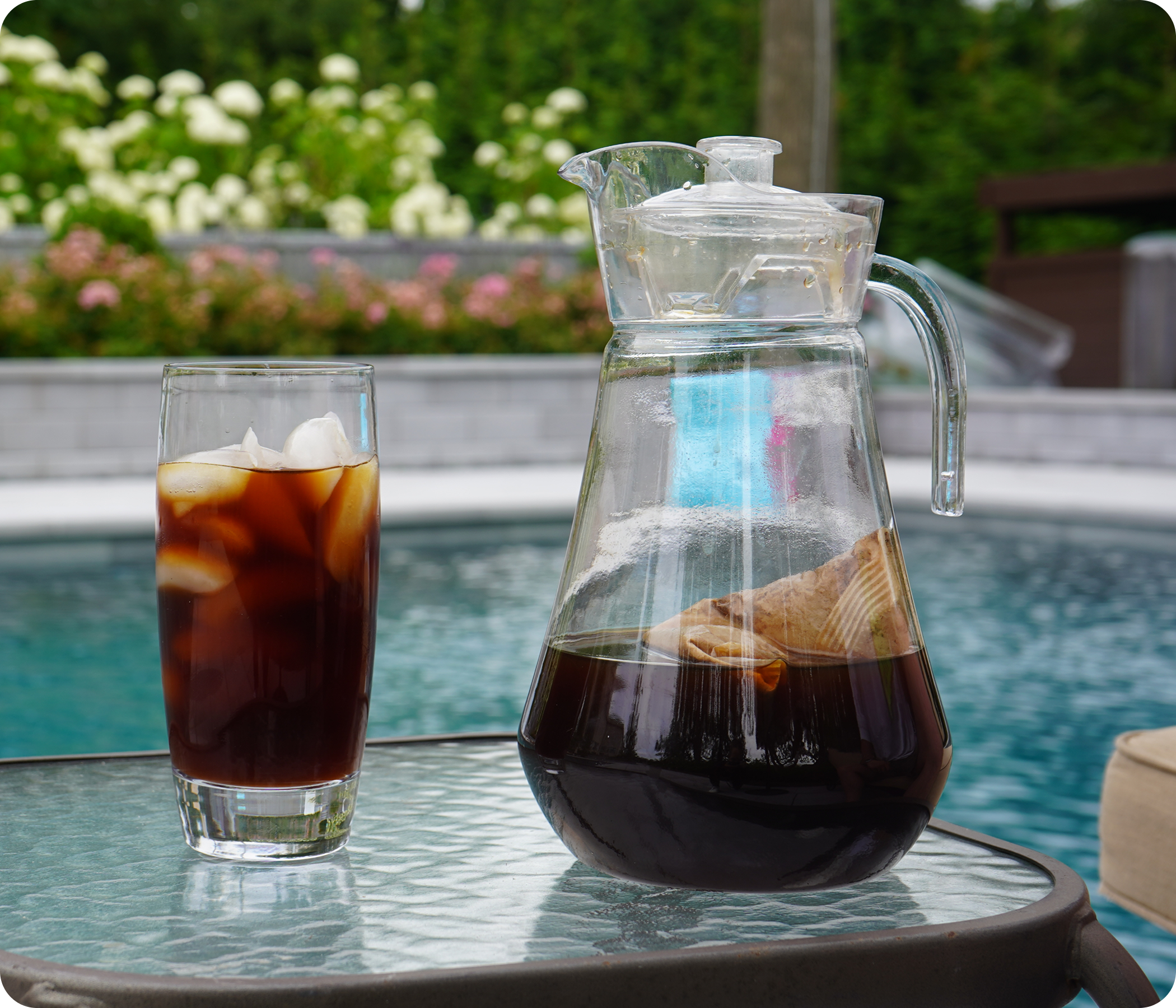 Iced coffee in a glass and a carafe on a table by a pool with a garden background