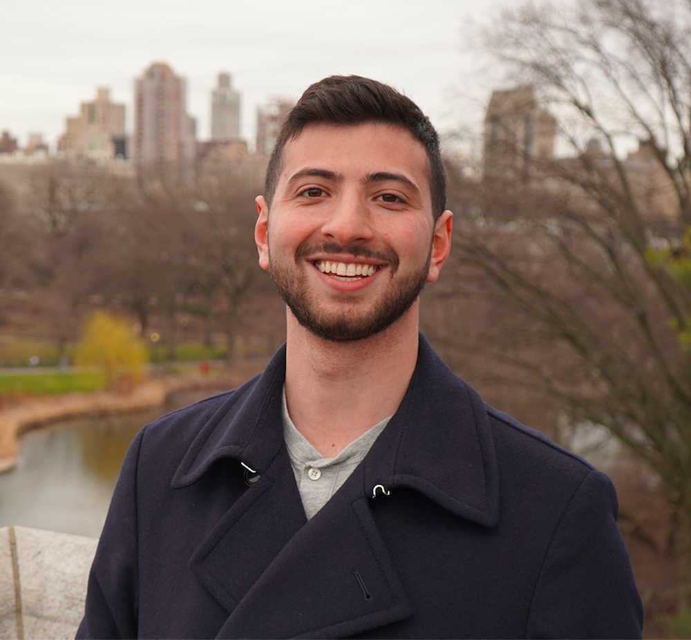 Man in a dark coat smiling with a cityscape and park in the background